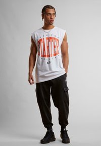 White sleeveless T-shirt with red graphic text "STARTER" and logo, paired with dark cargo-style sweatpants and black sneakers.
