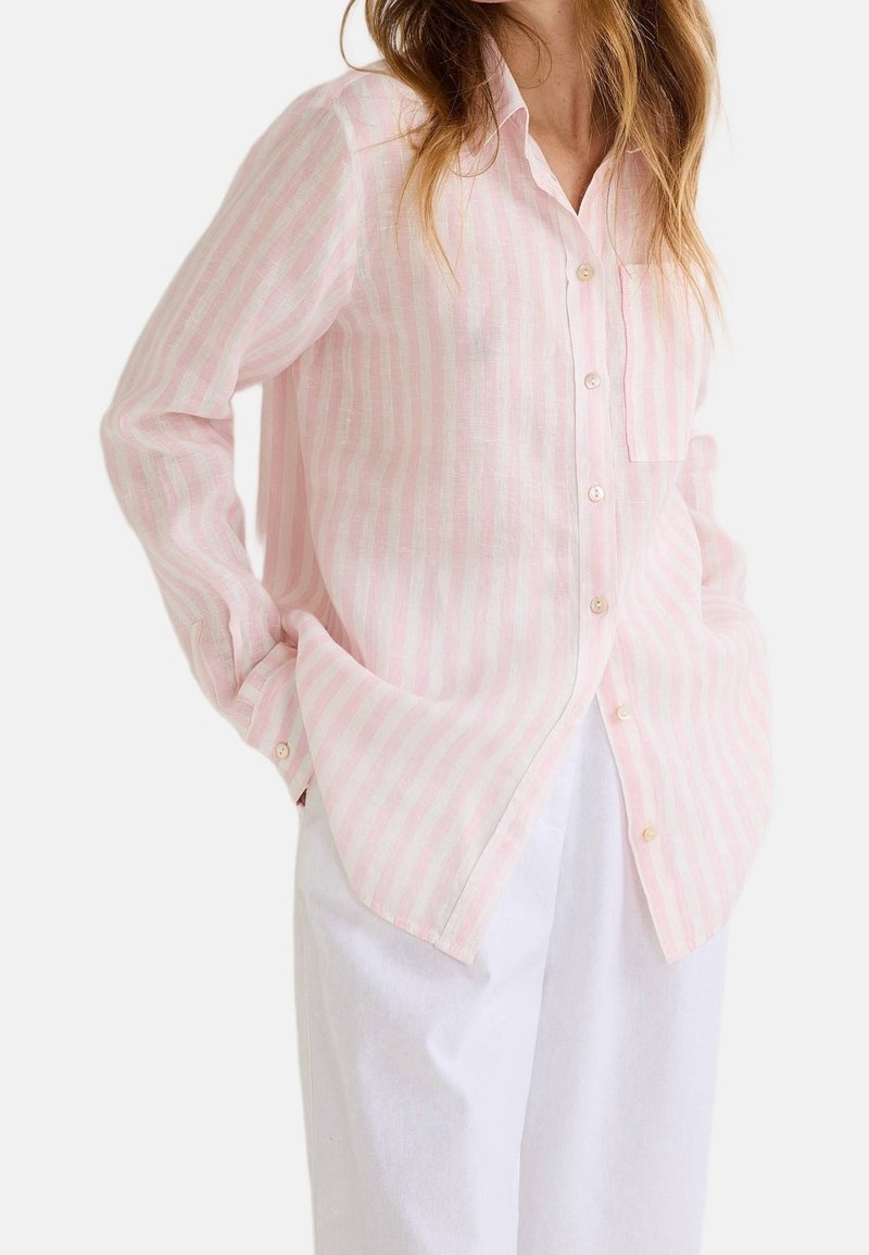 Femme portant une chemise rayée rose clair et blanche à boutons avec manches longues, associée à un pantalon blanc ample, les mains dans les poches.