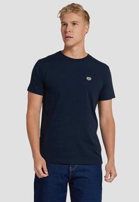 Camiseta de algodón azul marino con cuello redondo, mangas cortas y un pequeño logo texturizado en el lado izquierdo del pecho. Textura suave, ajuste casual.