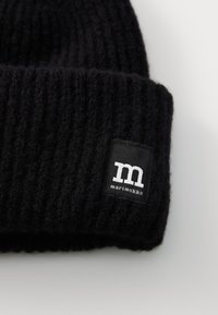 Čierna pletená čiapka s rebrovaním a prehnutým okrajom, na ktorom sa nachádza čierna nášivka s bielym písmenom „m“ a textom „marimekko.“