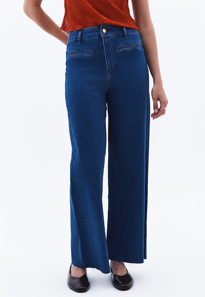 Modré džíny s vysokým pasem a širokými nohavicemi, mají dvě přední kapsy a raw lem. Obsahují jedno zlaté knoflíkové zapínání v pase.