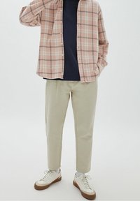 Camisa de cuadros rosa claro abotonada sobre una camiseta azul marino, combinada con pantalones beige ajustados y zapatillas blancas con detalles en marrón.