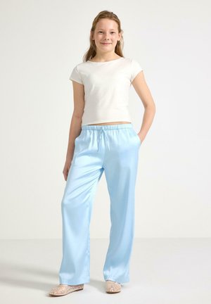 Nuori nainen seisoo kädet taskuissa, yllään valkoinen lyhythihainen paita, vaaleansiniset satiinihousut ja beiget kirjailtu ballerinat.