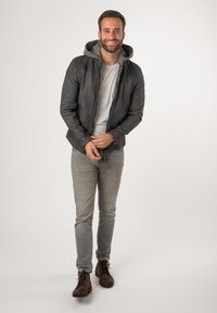 Giacca in pelle nera con cappuccio grigio, t-shirt grigia, jeans grigio chiaro e stivali marroni con lacci. Design semplice con dettagli di cucitura visibili.