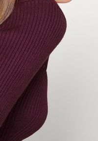 Primer plano lateral de una persona que lleva una prenda de manga larga acanalada de color púrpura oscuro sobre un fondo claro y sencillo.