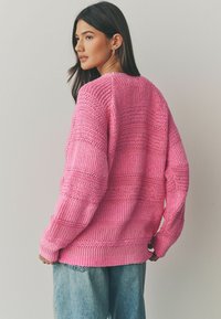 Rosa strikket sweater med et tekstureret design med vandrette striber og ribbede manchetter, parret med lyseblå denim jeans.