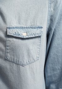 Lysblå, denim-lignende stofskjorte med en lomme med knaplukning, let krøllet tekstur og spids krave.