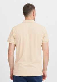 Beige kortermet polo-skjorte laget av teksturert stoff, med klassisk krage og rett kant, sett fra baksiden.