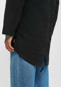Zwarte jas met een gebogen zoom, beschikt over zichtbare stiksels en een koord aan de onderkant, gecombineerd met lichtblauwe denim broek.
