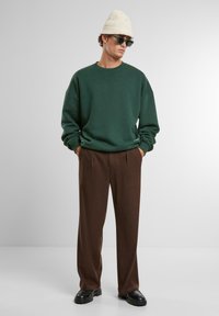 Tume rohelise värvi pullover, pruunid voltidega püksid, mustad kingad, beež kudumismüts ja rohelised prillid neutraalses taustas.