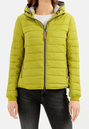Femme portant une veste matelassée vert citron avec une fermeture éclair à l'avant, des poches latérales zippées et une capuche, associée à un jean noir et une chemise blanche.