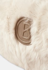 Flauschiger weißer Kunstpelzstoff mit einem strukturierten beigen Lederpatch, der einen geprägten Buchstaben "B" und gestickte Details aufweist.