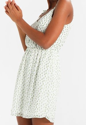 Biała sukienka bez rękawów z niewielkim zielonym nadrukiem kaktusów, z elastyczną talią i cienkimi ramiączkami, noszona przez osobę trzymającą ręce razem.