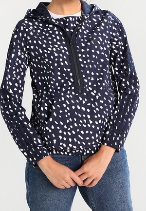 Polo à capuche bleu marine avec motif de points blancs irréguliers, fermeture éclair à l'avant, porté avec un jean bleu, mains de la personne jointes devant.