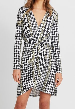 Czarna i biała sukienka kopertowa w kratę typu pepitka z długimi rękawami, zdobiona złotymi łańcuszkami i skośnym dołem. Gładka faktura tkaniny.