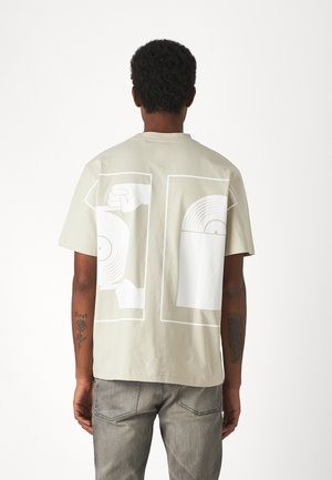T-shirt in cotone beige con grandi stampe grafiche bianche di dischi in vinile e mani sul retro. Maniche corte, vestibilità comoda, design minimale.