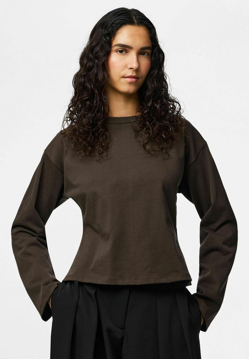 Jeune personne aux longs cheveux bouclés et foncés, portant un haut marron à manches longues et un pantalon noir, les mains dans les poches, sur un fond neutre.