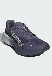Zapatilla de running de rendimiento en morado oscuro con parte superior de malla, acentos texturizados, gran logo "TERREX" y entresuela gris en contraste.