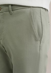 Pantalones de algodón verde claro con una textura suave, que presentan bolsillos laterales y un diseño de pierna recta, parcialmente cubiertos por un suéter blanco.
