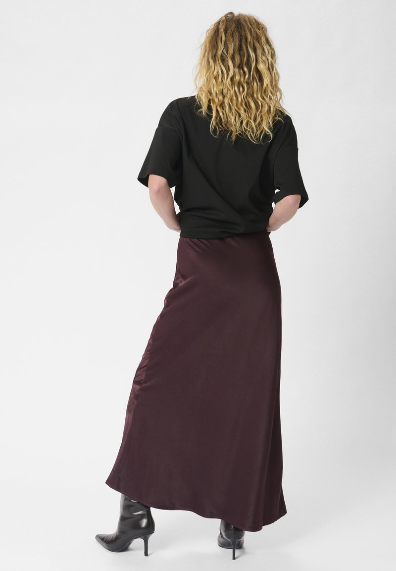 Burgunderfarbener Satin-Maxirock mit glatter Textur, kombiniert mit einem schwarzen, oversized Kurzarmoberteil und figurbetonten schwarzen Stiefeletten mit Absatz.