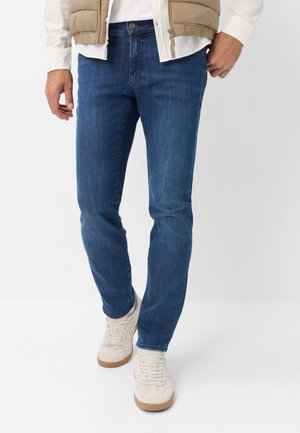 Uomo che indossa jeans blu slim fit, sneakers beige, camicia bianca e giacca imbottita beige, in piedi su sfondo bianco.