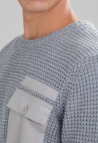 Grå stickad tröja med en strukturerad yta, försedd med en ljusgrå tygficka och knappstängning på vänster bröst.