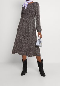 Schwarzes, floral gemustertes Midi-Kleid mit langen Ärmeln und gerafftem Taillenbund. Kombiniert mit einer kleinen, hellblauen Handtasche und schwarzen knielangen Stiefeln.