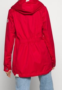 Červená nepromokavá bunda se staženým pasem, kapucí a manžetami na knoflíky. Hladká textura s viditelně odolnou tkaninou a logem na lemu.