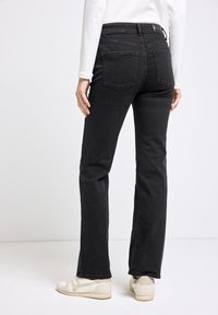 Jeans neri a zampa d'elefante con vita alta, dotati di due tasche posteriori e un patch in pelle sul retro, abbinati a sneakers chiare.