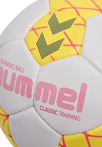 Vit träningsboll med gula och rosa detaljer som har en hexagonal design. Visar "Hummel Classic Training" i rosa text och linjer.