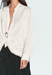 Blouse en satin blanc avec un décolleté en V profond, ornée d'un détail noué à l'avant et d'un accent doré. Manches longues et coupe décontractée.