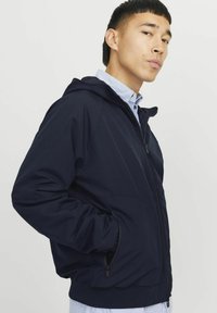 Veste zippée bleu marine avec capuche, présentant une texture lisse, des poches latérales et des poignets élastiques. Portée par-dessus une chemise à rayures blanches et bleues.