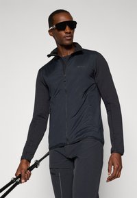 Homme portant une veste d'extérieur noire et des lunettes de soleil, tenant des bâtons de randonnée devant un fond clair.