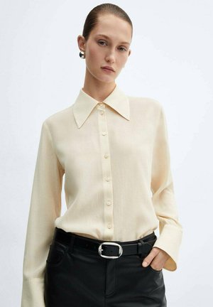 Blusa crema con un ampio colletto, bottoni sul davanti e maniche lunghe, abbinata a pantaloni neri e una cintura con fibbia d'argento.