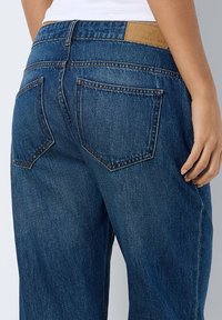 Jeans de ganga azul-escuro com um corte reto, bolsos traseiros proeminentes e um remendo em couro castanho na cintura. Detalhe de costura distintivo.