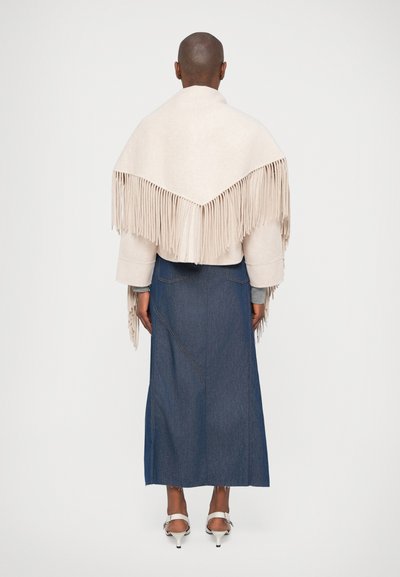 Beige sjal med fransar över en grå långärmad topp, kombinerat med en mörk denim maxikjol och vita klacksandaler, sedd bakifrån.