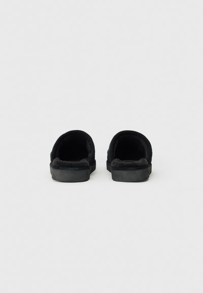 Zwarte instapslippers met een zachte, pluizige binnenkant en een vlakke rubberen zool. Minimalistisch ontwerp, zonder zichtbare hardware of patronen.