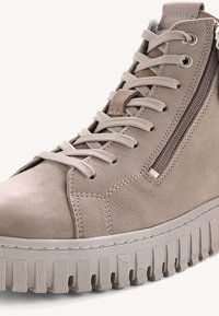 Beige korkeakartioinen lenkkitossu, joka on valmistettu mokasta ja jossa on kuvioitu kumipohja. Varustettu nauhoilla ja sivuvetoketjulla, joka helpottaa pääsyä.