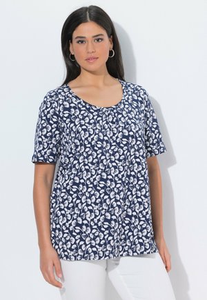 Kvinde iført marineblå kortærmet top med hvidt blomstermønster og hvide bukser, stående mod en ensfarvet lys baggrund.