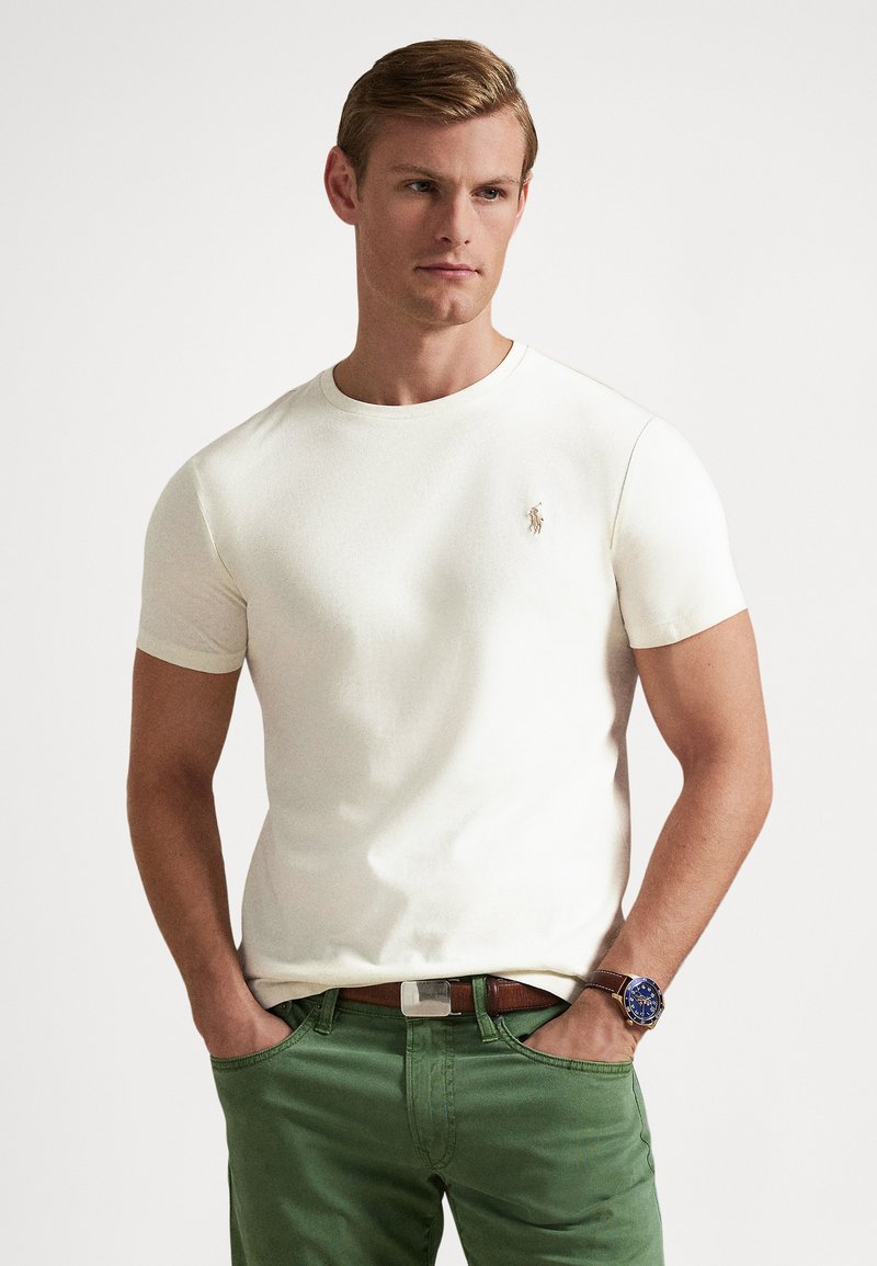 T-shirt blanc à manches courtes avec un col rond et un petit logo brodé. Porté avec un pantalon vert et une montre bleue avec un bracelet marron.