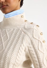 Creme kabelstrikket sweater med høj ribkant. Guldknapper pryder den ene skulder. En blå- og hvidstribet skjortekrave stikker frem nedenunder.
