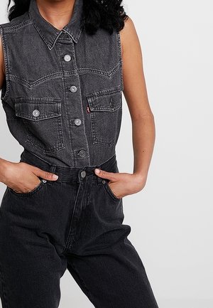 Bezrekawnikowa czarna koszula z denimu zapinana na guziki, z dwoma zapinanymi na guziki kieszeniami z przodu, noszona z wysokim stanem czarnych spodni z denimu, ręce w kieszeniach.