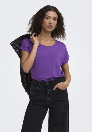 Woman with curly hair wearing a purple t-shirt and black jeans holds a black jacket over her shoulder against a plain background.