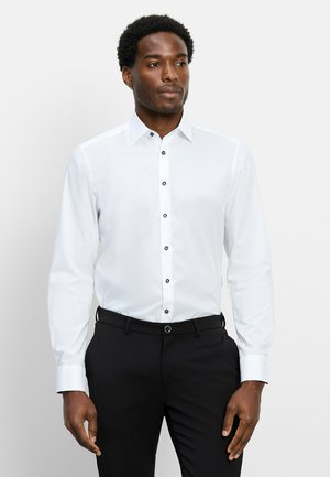 Mann trägt ein weißes langärmeliges Hemd mit Knöpfen und schwarze Hose, steht vor einem schlichten hellen Hintergrund.