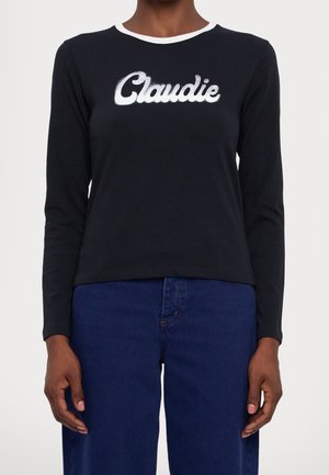 Zwart shirt met lange mouwen met de witte tekst "Claudie" op de borst, gecombineerd met een blauw jeans met hoge taille, gedragen door een staande persoon.
