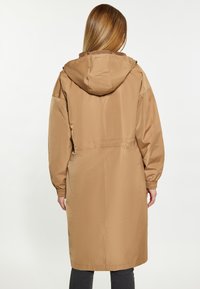 Lång, beige regnjacka med huva. Har elastiska ärmslut, dragsko i midjan och slät, lättviktsmaterial utan synliga knäppningar.