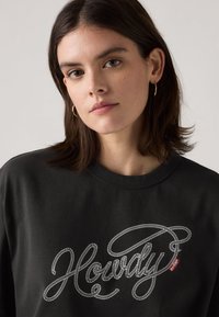 Sudadera negra hecha de un tejido suave que presenta un texto blanco en estilo cuerda que dice "Howdy" en el pecho, con una etiqueta de logo roja en el lateral.