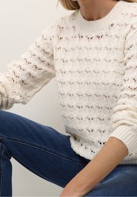 Pull en tricot couleur crème avec un motif de mailles texturé et ajouré. Présente des poignets côtelés et un col rond. Porté avec un jean en denim bleu.