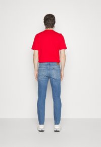 Camiseta roja de manga corta, jeans de mezclilla azul con lavado claro, vista trasera. Calzado son zapatillas blancas con detalles en negro.