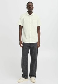 Camisa de botones de manga corta, color verde claro, con tejido texturizado y bolsillo en el pecho, combinada con jeans gris oscuro y zapatillas blancas.